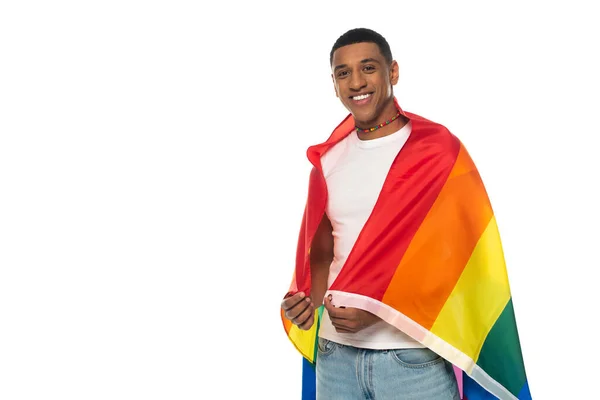 Joven afroamericano, envuelto en bandera lgbt, sonriendo a la cámara aislada en blanco - foto de stock