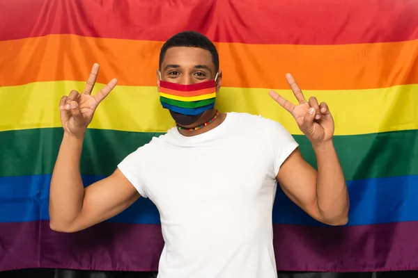 Homem americano africano em cores arco-íris máscara médica mostrando sinal de paz no fundo da bandeira lgbt — Fotografia de Stock