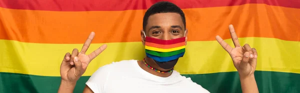 Homem americano africano em cores arco-íris máscara médica mostrando gesto de vitória no fundo da bandeira lgbt, bandeira — Fotografia de Stock