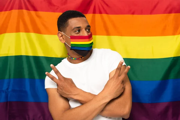 Hombre afroamericano con cuentas de colores arco iris y máscara médica, mostrando el gesto de victoria en el fondo de la bandera lgbt - foto de stock