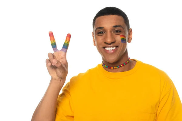 Homme afro-américain positif avec drapeau lgbt peint sur le visage montrant signe de paix isolé sur blanc — Photo de stock