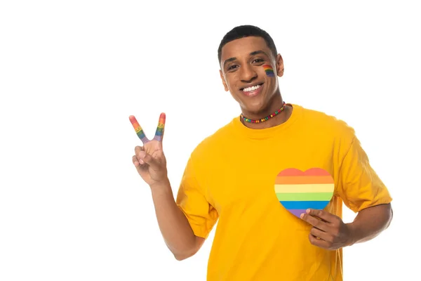 Homme afro-américain avec drapeau lgbt peint sur le visage, montrant signe de paix et coeur en papier dans des couleurs arc-en-ciel isolé sur blanc — Photo de stock