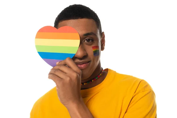 Homem americano africano com bandeira lgbt pintada no rosto, cobrindo o olho com cores do arco-íris coração de papel isolado no branco — Fotografia de Stock