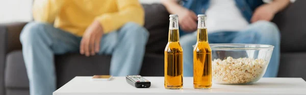 Bottles of beer, smartphone, tv remote and popcorn near friends sitting on blurred background, banner — Stock Photo