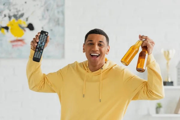 Sorridente afro-americano homem com TV remoto e garrafas de cerveja em comemorar pose no fundo borrado — Fotografia de Stock