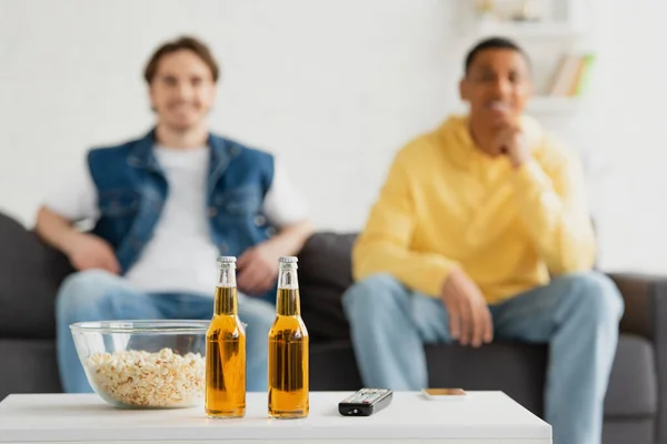 Botellas de cerveza, TV remoto y palomitas de maíz sobre fondo borroso con dos amigos interracial sentados en el sofá - foto de stock