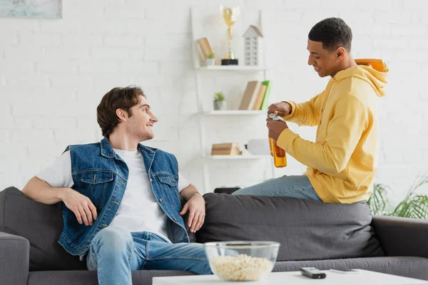 Jovem afro-americano abrindo garrafas de cerveja e descansando com o amigo na sala de estar — Fotografia de Stock