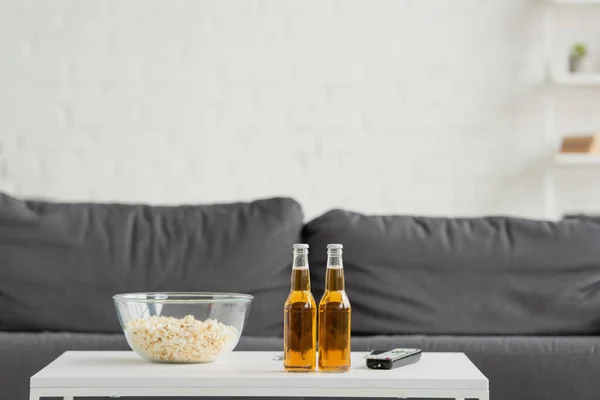 Vue de face de deux bouteilles de bière, pop-corn et télécommande TV sur fond flou avec canapé — Photo de stock