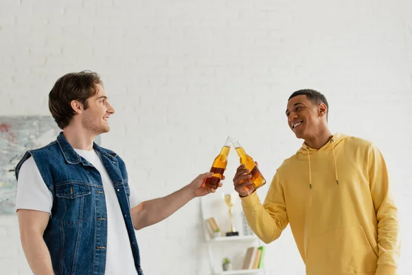 Feliz jovem interracial amigos clinking com garrafas de cerveja no loft moderno — Fotografia de Stock