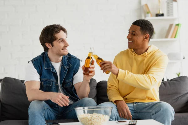 Glückliche junge interrassische Freunde, die im Wohnzimmer mit Bierflaschen klingeln — Stockfoto