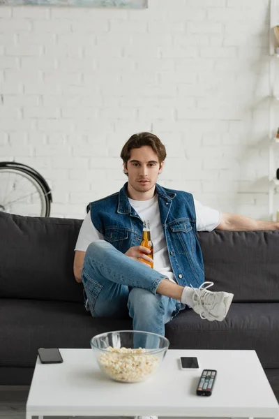 Giovane uomo premuroso seduto con birra sul divano vicino al tavolo con popcorn a casa — Foto stock