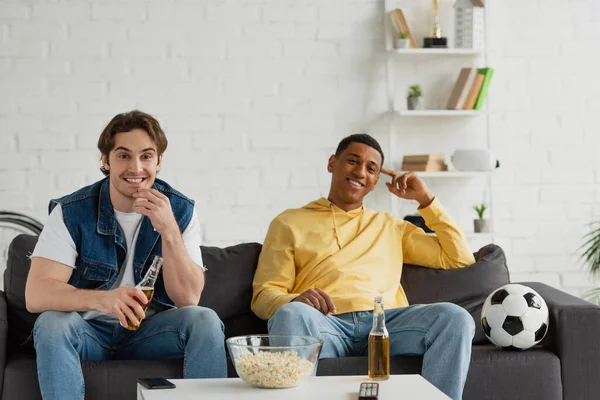 Два межрасовых футбольных болельщика сидят с пивом и попкорном и смотрят игру дома — стоковое фото