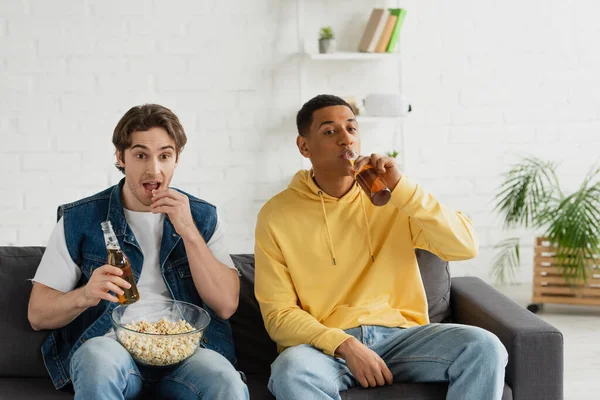 Homme afro-américain avec un ami boire de la bière et manger du pop-corn, assis sur le canapé à la maison — Photo de stock