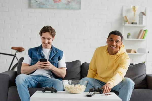 KYIV, UKRAINE - MARCH 22, 2021: young man resting together with african american friend and looking at smartphone near table with joysticks — Stock Photo