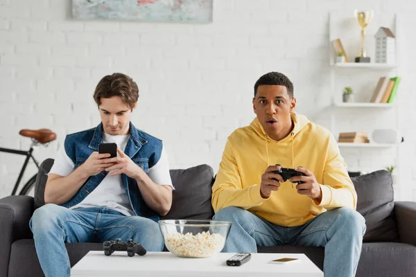 KYIV, UKRAINE - MARCH 22, 2021: young man looking at smartphone near african american friend playing with joystick at home — Stock Photo