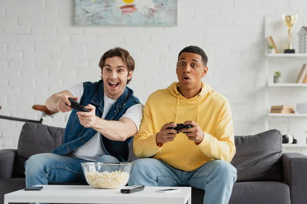 KYIV, UKRAINE - MARCH 22, 2021: interracial friends emotionally playing video game with joysticks — Stock Photo