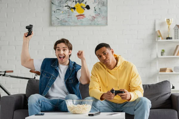 KYIV, UCRANIA - 22 de marzo de 2021: jóvenes amigos interracial jugando videojuegos con joysticks en la moderna sala de estar - foto de stock