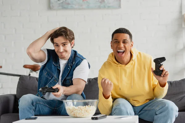 KYIV, UKRAINE - MARCH 22, 2021: interracial friends emotionally playing video game with joysticks in arms in living room — Stock Photo