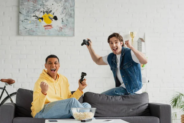 KYIV, UKRAINE - MARCH 22, 2021: modern interracial friends emotionally playing video game with joysticks at home — Stock Photo