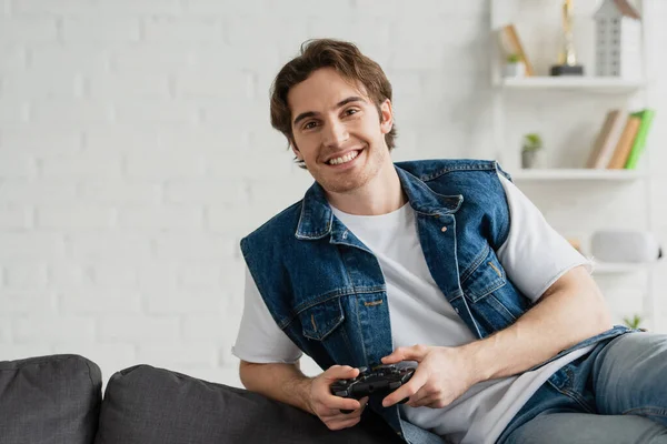 KYIV, UKRAINE - MARCH 22, 2021: young smiling man sitting in relax pose on couch with joystick in modern room — Stock Photo