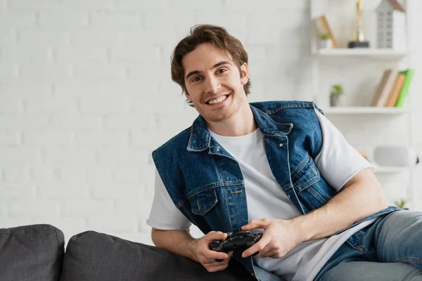 KYIV, UCRANIA - 22 de marzo de 2021: joven sonriente sentado en pose de relax en el sofá con joystick en la habitación moderna - foto de stock