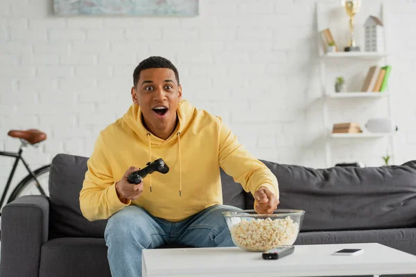 KYIV, UKRAINE - 22 MARS 2021 : homme afro-américain excité assis avec joystick sur le canapé dans le salon — Photo de stock