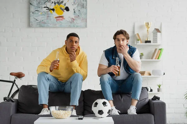 QUIIV, UCRÂNIA - MARÇO 22, 2021: amigos inter-raciais assistindo jogo de futebol com garrafas de cerveja e pipocas na sala de estar — Fotografia de Stock