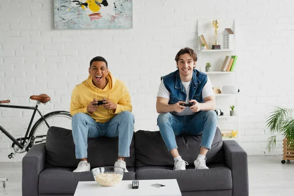 KYIV, UCRANIA - 22 de marzo de 2021: amigos interracial que se concentran en el videojuego con palillos y palomitas de maíz en la sala de estar - foto de stock