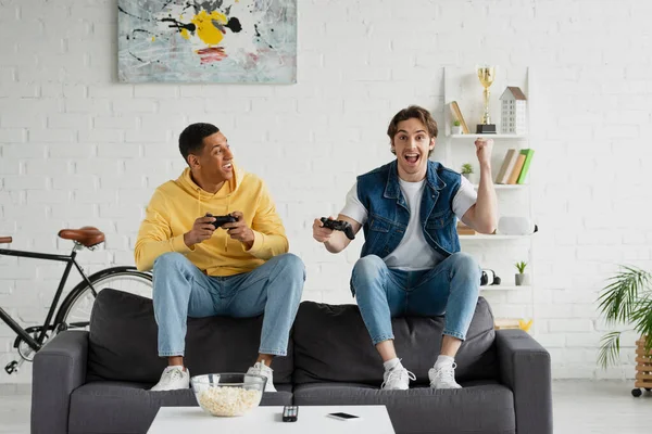 KYIV, UCRANIA - 22 de marzo de 2021: un joven hipster jugando un videojuego con un amigo afroamericano en un loft moderno - foto de stock