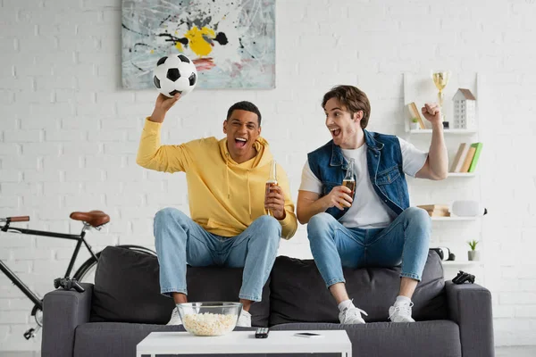 KYIV, UKRAINE - MARCH 22, 2021: young interracial friends celebrating victory with beer and football in modern loft — Stock Photo