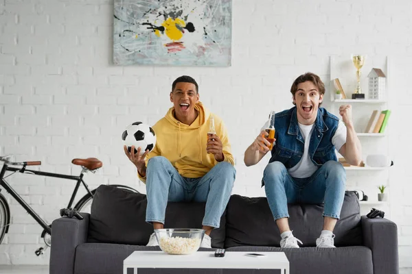 KYIV, UKRAINE - MARCH 22, 2021: young interracial football fans sitting on couch and celebrating triumph with beer and popcorn in modern loft — Stock Photo