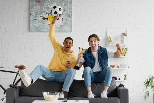 KYIV, UKRAINE - 22 MARS 2021 : joyeux supporters de football interracial assis sur le canapé et célébrant la victoire avec de la bière et une coupe de triomphe dans le loft — Photo de stock