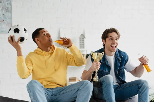 Fans de football interracial assis sur le canapé et boire de la bière avec tasse de triomphe dans le loft moderne — Photo de stock