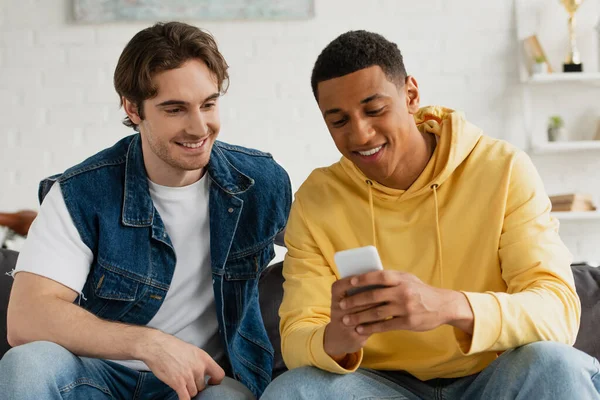 Interracial friends watching at smartphone together sitting on couch in living room — Stock Photo