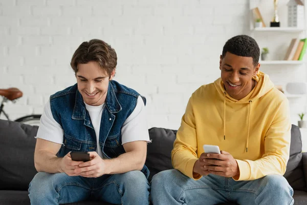 Smiling interracial friends sitting on couch and texting messages with smartphones in modern loft — Stock Photo