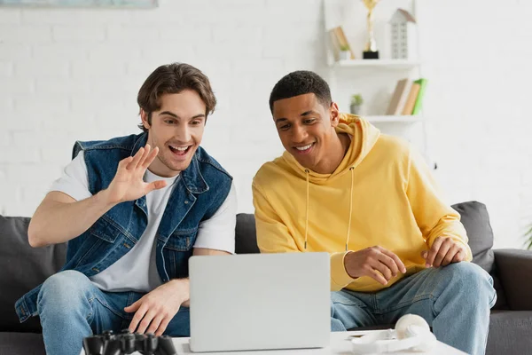 Amigos interracial felices sentados juntos en el sofá y haciendo videollamada con computadora portátil en la sala de estar - foto de stock