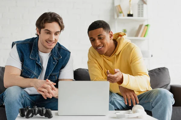 KYIV, UKRAINE - MARCH 22, 2021: interracial friends sitting together on couch in front of laptop in living room — Stock Photo