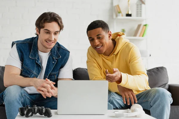 KYIV, UCRANIA - 22 de marzo de 2021: amigos interracial sentados juntos en el sofá frente a la computadora portátil en la sala de estar - foto de stock