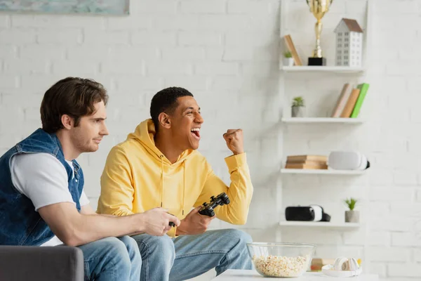 KIEW, UKRAINE - 22. MÄRZ 2021: Junge interrassische Freunde sitzen mit Steuerknüppeln am Tisch mit Popcorn und Kopfhörern im modernen Loft — Stockfoto