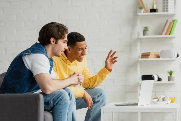 Fröhliche interrassische Freunde, die zusammen sitzen und am Laptop im modernen Wohnzimmer zusehen — Stockfoto