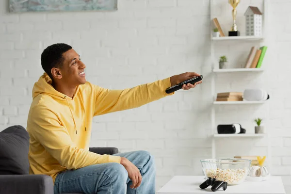 KYIV, UKRAINE - MARCH 22, 2021: young african american man with TV remote controller sitting on couch with popcorn, joystick and headphones in modern loft — Stock Photo