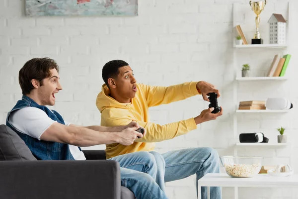 KYIV, UKRAINE - MARCH 22, 2021: joyful interracial friends sitting together on couch and playing video game with joysticks in modern loft — Stock Photo