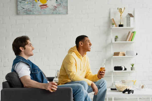 QUIIV, UCRÂNIA - MARÇO 22, 2021: amigos inter-raciais sentados no sofá, bebendo cerveja perto da mesa com pipoca, joystick, TV remota e fones de ouvido — Fotografia de Stock