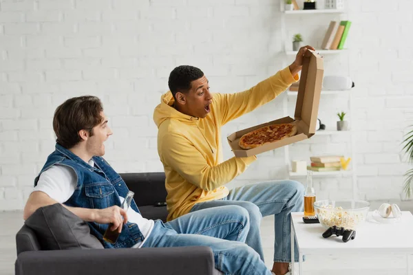 KYIV, UKRAINE - 22 MARS 2021 : Des amis interraciaux se réjouissent d'une pizza assise sur un canapé avec des bouteilles de bière dans un loft moderne — Photo de stock