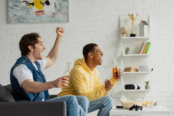 KYIV, UKRAINE - MARCH 22, 2021: interracial friends sitting on couch with bottles of beer and emotionally watching tv in living room — Stock Photo