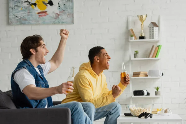 KYIV, UCRANIA - 22 de marzo de 2021: amigos interracial sentados en el sofá con botellas de cerveza y viendo emocionalmente la televisión en la sala de estar - foto de stock