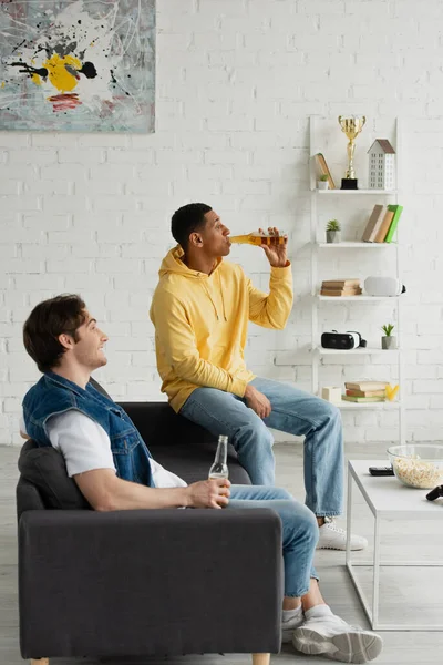 Side view of interracial friends resting on couch and drinking beer together in modern living room — Stock Photo