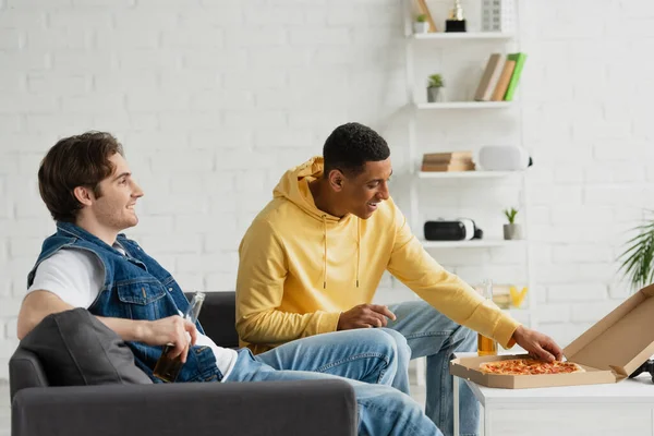 Interracial friends resting on couch and enjoying pizza with beer together in modern living room — Stock Photo