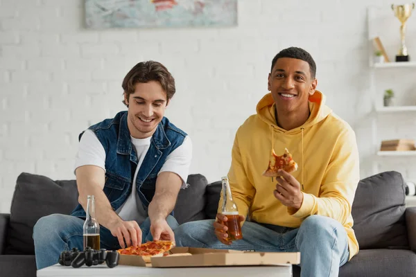 KYIV, UKRAINE - MARCH 22, 2021: interracial friends sitting on couch with bottles of beer and eating pizza in living room — Stock Photo