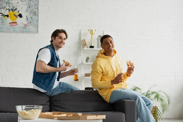 Young interracial friends enjoying pizza and beer on couch in modern living room — Stock Photo